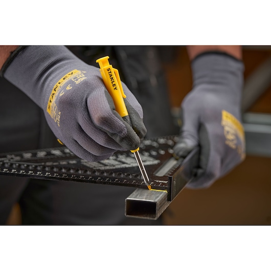 Gloved hands use a yellow STANLEY marking tool with ruler on a metal tube for precise measurement and marking.