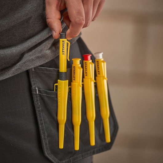 A hand holds a yellow STANLEY tool from a pocket with four yellow STANLEY marking tools clipped to work trousers.