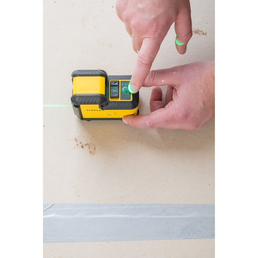 STANLEY STHT77594-1 laser level being operated, close-up of hand pressing power button, green laser line visible.