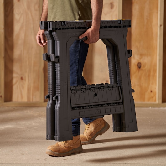 Photo of STANLEY STST1-70713 folding sawhorse being carried by a person in a workshop setting.