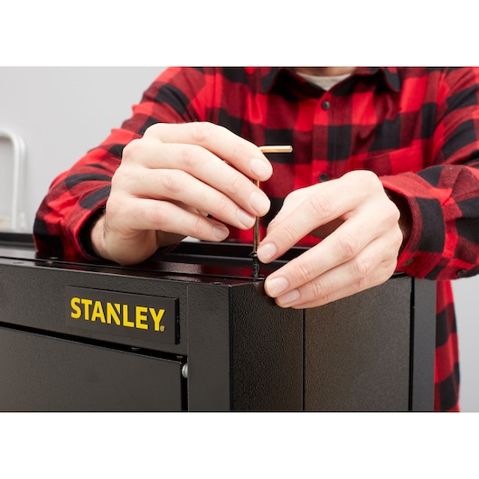 Photo of hands assembling STANLEY STST97599-1 tool chest with hex key, close-up view of logo and screw.